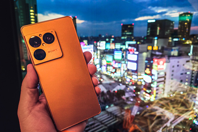 渋谷スクランブル交差点の夜景を背景にXperia 1 VIII ネイティブゴールドを手に持ってBluetooth通信テストを行った様子